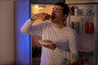 © Pixel-Shot - Hungry young man eating croissant near open fridge in kitchen at night