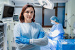 © InsideCreativeHouse - Portrait of female doctor looking at camera in operating room, blurred background with surgeons performing operation under surgical lights, professional healthcare surgery