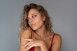© Марина Демешко - Sensual portrait of young attractive blonde woman with voluminous wavy hair, piercings and red lace bra, touching her shoulder and looking aside on grey background