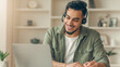 © Prostock-studio - A young man sits at a desk in a bright room with a laptop and headset. He smiles while taking notes and participating in an online learning session. The scene shows focus and dedication.