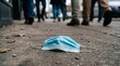 © HoneyStock - Discarded blue face mask on urban sidewalk with pedestrians walking by