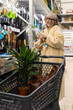 © Ivan Traimak - Happy senior woman shopping for gardening supplies, holding tools and pushing a cart
