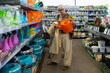 © Ivan Traimak - Senior woman shopping for gardening supplies, holding an orange watering can in a store aisle