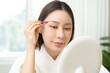 © KMPZZZ - Hair tweezer depilation concept. Beautiful asian young woman plucking eyebrows with tweezers looking at mirror. Female removal, epilation or facial, treatment for skincare, cleaning and cosmetic tool.