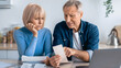 © Prostock-studio - A man and a woman sit at a kitchen table looking at receipts. The woman appears concerned while the man points to a receipt, suggesting they are discussing household expenses.