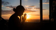 © Sabiq - A woman prays while looking out the window at a serene sunset over the landscape.