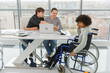 © Юлия Завалишина - Teamwork brainstorm. Diverse multiracial colleagues woman in chair for people with disability working together. Group of people sitting in creative office discussing business. Young professional team