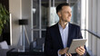 © fizkes - Confident and successful businessman stands by office window, smiling, reflecting on creative ideas, reviewing project updates on digital tablet. Strategic thinking, business decisions, technology