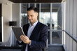 © fizkes - Focused businessman stands in office hallway with tablet, managing business tasks, reads information, review digital data, consulting corporate client via email, staying connected through technology
