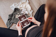 © Katrin_Primak - Close up of a pregnant woman holding ultrasound images of her baby next to tiny clothes on a sofa