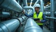 © miss irine - Engineer with tablet checks pipes in industrial facility. Man in hard hat and vest inspects complex piping system. Professional works on heavy machinery controls.