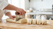 © CK Studio - Close-up of baker's hands using a metal scraper to portion fresh pastry dough on a floured wooden board in a commercial bakery.