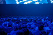 © Anton Gvozdikov - Large audience watching presentation in dark blue conference hall, corporate event atmosphere, focused attention and anticipation.