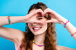 © deagreez - Smiling young woman making a heart hand gesture over eye with colorful beads and bracelets on bright blue background expressing love joy and playful portrait