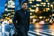 © whitestorm - Mature businessman walking in a city at night, carrying a leather bag, with blurred city lights in the background.