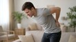 © AMAR - Young Man Suffering from Acute Lower Back Pain Standing in Living Room and Holding Spine in Discomfort