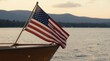 © Raisa - American flag waves on a wooden boat sailing on calm water at sunset. Mountains and trees form a scenic background on the horizon. This represents patriotism and freedom.