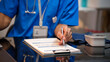 © NanSan - Close-up of healthcare worker in blue scrubs filling out medical forms on clipboard at desk, with stethoscope and badge visible, clinical office setting.