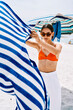 © Studio Marmellata - A young woman in sunglasses and an orange bikini top holds a blue and white striped towel flowing in the breeze on a sunny beach with umbrellas.