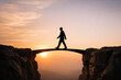 © DailyStock - Silhouette of a businessman walking across a narrow stone bridge connecting two rocky cliffs during a soft golden sunset, conceptual business