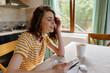 © SHOTPRIME STUDIO - Young woman with curly hair sits at a sunlit kitchen table, smiling softly while tapping a tablet in a striped yellow shirt.