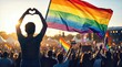 © Mariya Sorvacheva - Individual holding rainbow flag and making heart gesture at LGBTQ Pride Parade with crowd and colorful flags in the background during a vibrant outdoor celebration