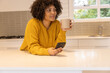 © wavebreak3 - Woman leaning on kitchen island in mustard sweater, holding beige mug and smartphone, gazing