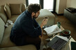 © TrueFrame Collective - High angle view of middle aged Caucasian man drinking coffee while reviewing documents beside laptop at home, showing remote work stress