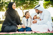 © oneinchpunch - Arabian family in traditional dress smiling together on a picnic blanket in a sunny green park, parents and children enjoying leisure, bonding and relaxation outdoors