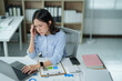 © MINAE - Stressed asian business woman experiencing a severe headache and burnout while working on her laptop in a modern office, overwhelmed by financial data and paperwork