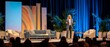 © Agung - A confident woman in a blue blazer speaks on a stage with modern furniture and colorful backdrops, addressing an audience