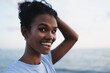© SISMIZ - Young woman with dark curly hair tied up, smiling and looking at the camera by the sea