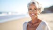 © inayatul - Smiling mature woman with short grey hair, joyful expression, standing on a sandy beach with ocean waves in background, enjoying summer vacation, healthy aging concept