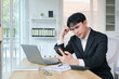 © amnaj - Young Asian businessman sitting at office desk, looking worried and frustrated while holding a phone and processing information