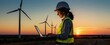 © Pete - Smiling woman in hard hat and safety vest checks laptop by wind turbines at sunset. She works on renewable energy project. Woman inspects wind farm operation.