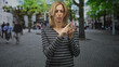 © Krakenimages.com - Blonde woman in striped top pressing her index finger with the other hand while grimacing in a cobbled street plaza lined with trees; concern focus.
