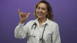 © Krakenimages.com - Middle aged hispanic woman doctor wearing a white shirt and stethoscope smiles and raises hand in purple studio; compassion.