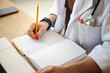 © Studio Marmellata - A medical professional in a white coat writes notes in a journal with a pencil, a stethoscope around their neck, and a laptop nearby.