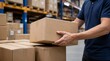 © Rhony - A logistics worker hand lifts a sealed cardboard box from a pile of boxes in a bright warehouse environment.