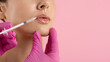 © Anastasiya - Closeup of young woman receiving hyaluronic acid lip filler injection by doctor hands in pink gloves, lip augmentation cosmetic procedure on pink background