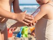 © Marian - Primer plano de un niño aplicando protector solar a su hermano menor en una playa con juguetes difuminados al fondo.
