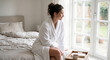 © Chari - A woman in a white bathrobe sits on a bed next to a window, looking outside with a cup of tea on a tray.
