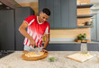 © wavebreak3 - Slicing man in red-white shirt using pizza cutter on freshly baked pizza on marble counter, basil