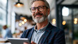 © Denis Yevtekhov - Happy middle-aged male executive in a modern office looking away thoughtfully while holding a tablet device with a pleasant expression, defocused bright open workspace in