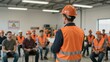 © mihrzn - A construction worker in an orange hard hat and safety vest leads a training session, with other workers attentively listening in a workshop setting.