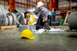 © Stella - Yellow safety helmet on ground with an industrial engineer injured leg in an accident at worksite, lying on factory floor. Injured worker feels in pain, need help. Senior supervisor tries to help him.