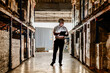 © ultramansk - Silhouette of a supply chain coordinator inspecting high pallet racks and walking towards a shipping container dock inside a commercial logistics terminal.