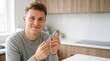© BETTOVERSE - Portrait of a smiling young man holding an insulin pen or medical injection device in a bright, modern kitchen, representing diabetes management or weight loss treatment