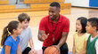 © Aurora Aesthetics - african american male physical education teacher coach elementary kids gym fitness sports athletic teacher leading playful warmup activity with diverse young students in colorful indoor gymnasium