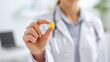 © KotBaton - Close-up of healthcare professional holding yellow softgel vitamin capsule between fingers in modern clinic setting, focus on supplement, blurred medical background, healthcare and wellness concept